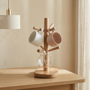 Puede incluir: Un árbol de tazas de madera con una base redonda y clavijas para colgar tazas. Se muestran tres tazas: una blanca, una rosa y una taza de vidrio transparente. El árbol está hecho de madera clara y está sobre una mesa de madera.