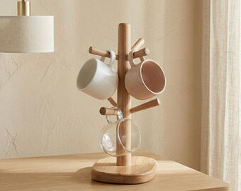 May include: A wooden mug tree with a round base and multiple pegs to hold mugs. Three mugs are displayed: a white, a pink, and a clear glass mug. The tree is made of light-colored wood and sits on a wooden table.