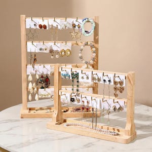 May include: Two wooden earring display stands showcasing various earrings, bracelets, and necklaces. The earrings are displayed on white cards, and the stands are made of light-colored wood. The jewelry includes star, pearl, and floral designs.