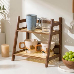 Puede incluir: Una estanter&iacute;a de madera de dos niveles con una superficie tejida de color amarillo claro. El estante superior contiene una taza azul y un vaso transparente. El estante inferior muestra dos tazas con un dise&ntilde;o de paisaje. Tambi&eacute;n se ven una vela y un cuenco de uvas.