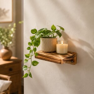 May include: A small, rectangular, dark-stained wooden wall shelf mounted with visible metal hardware. On the shelf sits a cream-coloured, vertically ribbed ceramic pot containing a trailing green pothos plant and a clear glass candle.