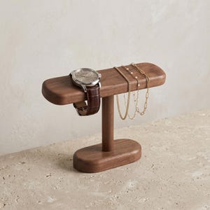 Peut inclure: Un présentoir en bois marron foncé pour montres et bijoux. Le présentoir comprend une barre horizontale pour une montre avec un bracelet en cuir marron. Plusieurs colliers dorés sont suspendus à la barre.