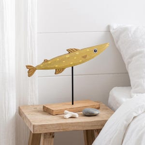 Puede incluir: Escultura de pez de madera amarilla y marrón, montada sobre un soporte negro y una base de madera. El pez tiene un diseño moteado y aletas. La escultura se exhibe sobre una mesa de madera rústica, con un hueso pequeño y una piedra gris.