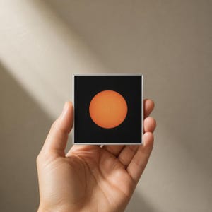 Op de afbeelding: Een vierkant object met een zwarte achtergrond en een gecentreerde, feloranje cirkelvormige zongrafiek. Het ontwerp bevat een dunne zilverkleurige rand rondom het vierkant, wat zorgt voor een strakke en gedefinieerde afwerking.