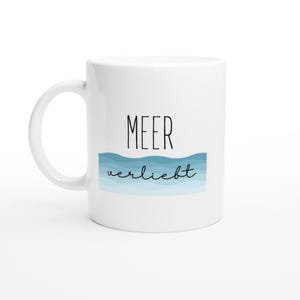 Peut inclure: Un mug en céramique blanche avec le texte MEER en police sans-serif noire au-dessus d'un motif ondulé bleu. Sous le motif, le mot verliebt est écrit en écriture cursive noire. Le mug possède une anse incurvée sur le côté gauche.