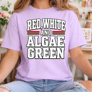 Peut inclure: Un t-shirt à manches courtes violet clair avec le texte RED, WHITE AND ALGAE GREEN en lettres capitales noires, grasses et usées. Le motif comprend des bandes horizontales rouges et vertes situées entre les lignes de texte.