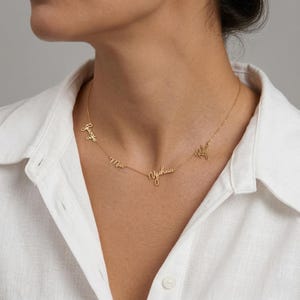 Può includere: Una delicata collana d'oro con più nomi in corsivo. I nomi sono collegati da una sottile catena. La collana è indossata su una camicia bianca.