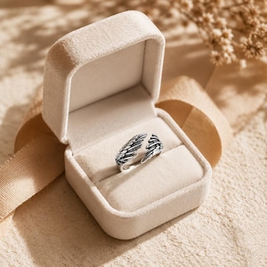 Op de afbeelding: Een open ringdoosje van beige fluweel met een zilverkleurige metalen ring in de vorm van twee overlappende engelvleugels. De ring heeft geoxideerde details in de groeven van de veren om de textuur van het vleugelontwerp te benadrukken.