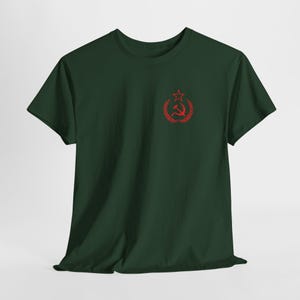 May include: Dark green t-shirt featuring a red emblem of a star, hammer, and sickle within a wreath. The short-sleeved shirt is made of a soft material and has a crew neck. The emblem is located on the upper left chest area.