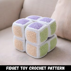 Può includere: Un giocattolo fidget all'uncinetto fatto a mano a forma di cubo. Il giocattolo è composto da cubi più piccoli nei colori pastello: lavanda, verde chiaro e pesca, con bordi bianchi. Il testo "FIDGET TOY CROCHET PATTERN" è in basso.