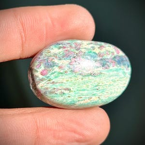 May include: An oval gemstone with a mix of green, pink, and white hues. The polished stone has a natural appearance. The gemstone is held between two fingers. The stone's dimensions are approximately 3 cm x 2 cm.