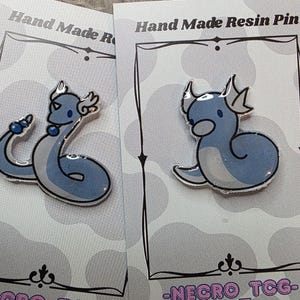 May include: Two handmade resin pins featuring a blue and white dragon design. Each pin is mounted on a gray card with the text "Hand Made Resin Pin" and "-NECRO_TCG-". The pins are on a fluffy, gray surface.