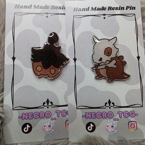 May include: Two resin pins on cards. One pin is a brown and orange pumpkin-like character with a silver outline. The other pin is a brown and white cartoon character with a bone-like head. The cards read "Hand Made Resin Pin" and "-NECRO_TCG-".