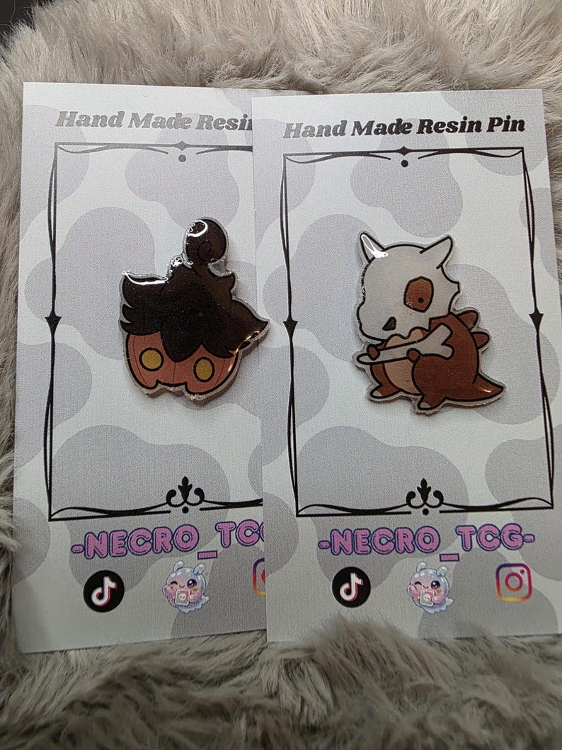 May include: Two resin pins on cards. One pin is a brown and orange pumpkin-like character with a silver outline. The other pin is a brown and white cartoon character with a bone-like head. The cards read "Hand Made Resin Pin" and "-NECRO_TCG-".