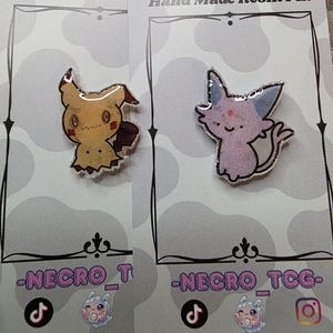 May include: Two handmade resin pins on display. One pin features a yellow cartoon character with a brown tail and a jagged bottom. The other pin is a pink cartoon cat with blue ears. Both pins are on gray cards with the text "Hand Made Resin Pin".