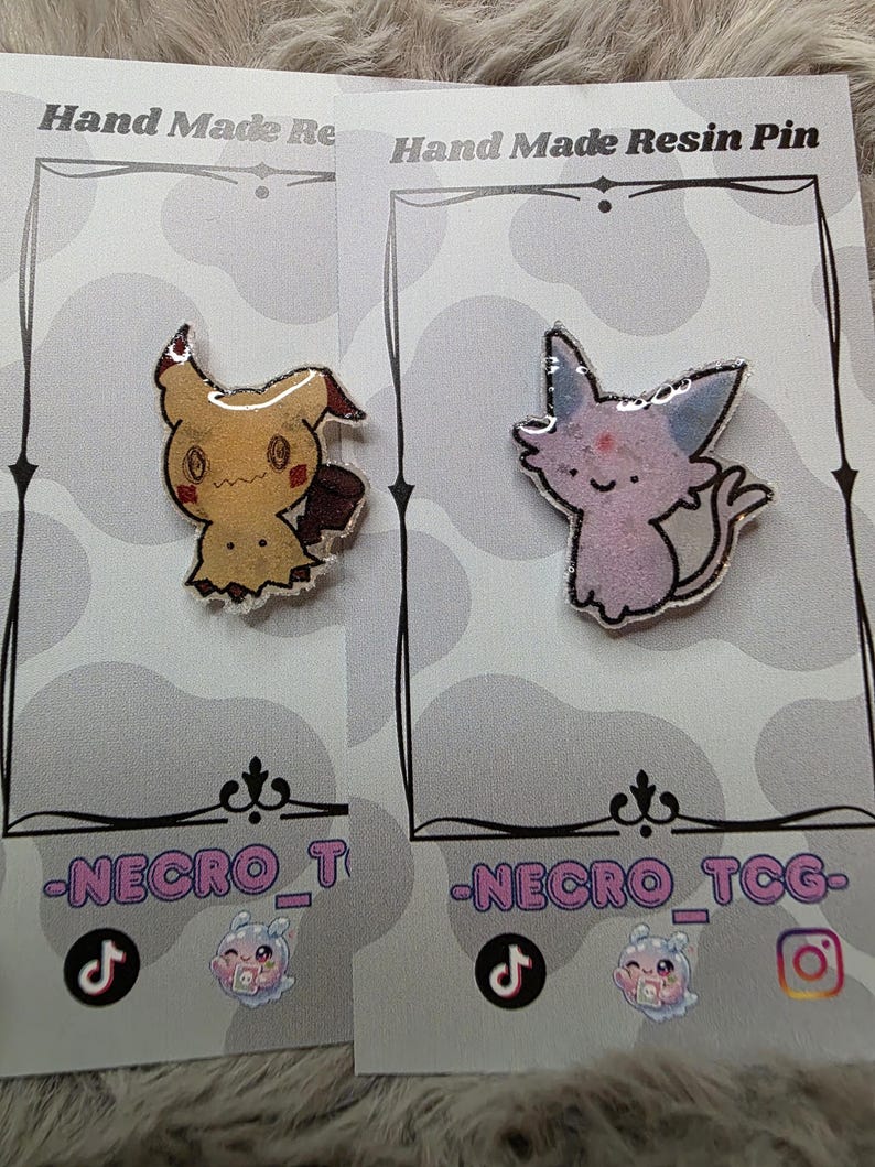 May include: Two handmade resin pins on display. One pin features a yellow cartoon character with a brown tail and a jagged bottom. The other pin is a pink cartoon cat with blue ears. Both pins are on gray cards with the text "Hand Made Resin Pin".
