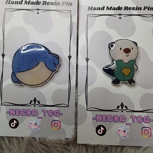 May include: Two resin pins on display. One pin features a blue whale design, and the other depicts an otter wearing a green shirt. Both pins are on cards that read "Hand Made Resin Pin" and include social media handles.