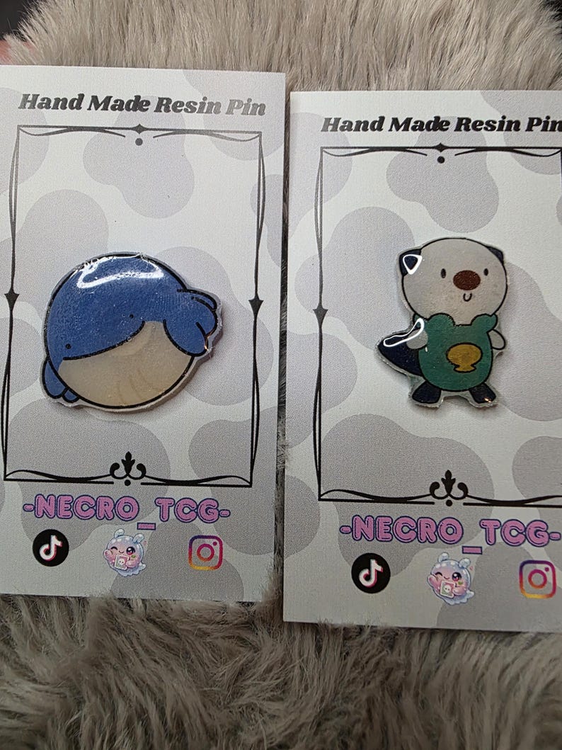 May include: Two resin pins on display. One pin features a blue whale design, and the other depicts an otter wearing a green shirt. Both pins are on cards that read "Hand Made Resin Pin" and include social media handles.