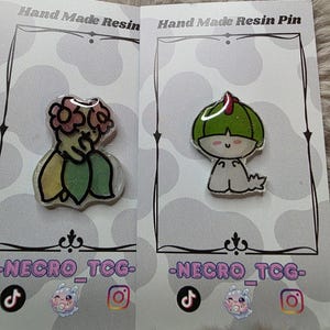 May include: Two resin pins on display. One pin features a green and yellow floral design, the other a green character with a red accent. Both pins are on cards with the text "Hand Made Resin Pin" and "-NECRO_TCG-".