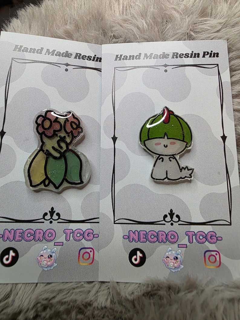 May include: Two resin pins on display. One pin features a green and yellow floral design, the other a green character with a red accent. Both pins are on cards with the text "Hand Made Resin Pin" and "-NECRO_TCG-".