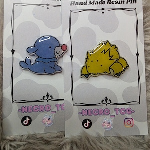 May include: Two handmade resin pins on cards. One pin is a blue seal with a red nose, the other is a yellow, spiky creature. The cards have the text "Hand Made Resin Pin" and "-NECRO_TCG-".