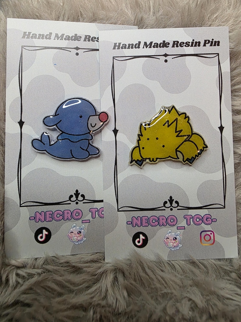 May include: Two handmade resin pins on cards. One pin is a blue seal with a red nose, the other is a yellow, spiky creature. The cards have the text "Hand Made Resin Pin" and "-NECRO_TCG-".