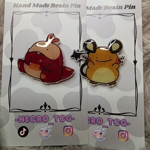 May include: Two resin pins on cards. One pin is a red and brown cartoon creature. The other is a yellow and brown cartoon mouse with black ears. The cards have the text "Hand Made Resin Pin" and "-NECRO_TCG-".