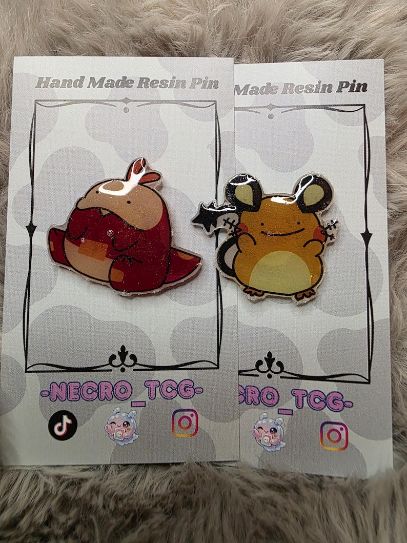 May include: Two resin pins on cards. One pin is a red and brown cartoon creature. The other is a yellow and brown cartoon mouse with black ears. The cards have the text "Hand Made Resin Pin" and "-NECRO_TCG-".