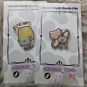 May include: Two handmade resin pins on display. One pin features a yellow design with the text "I'M YOU GOD". The other pin has a pink cat and bunny design. Both pins are on white cards with black borders.