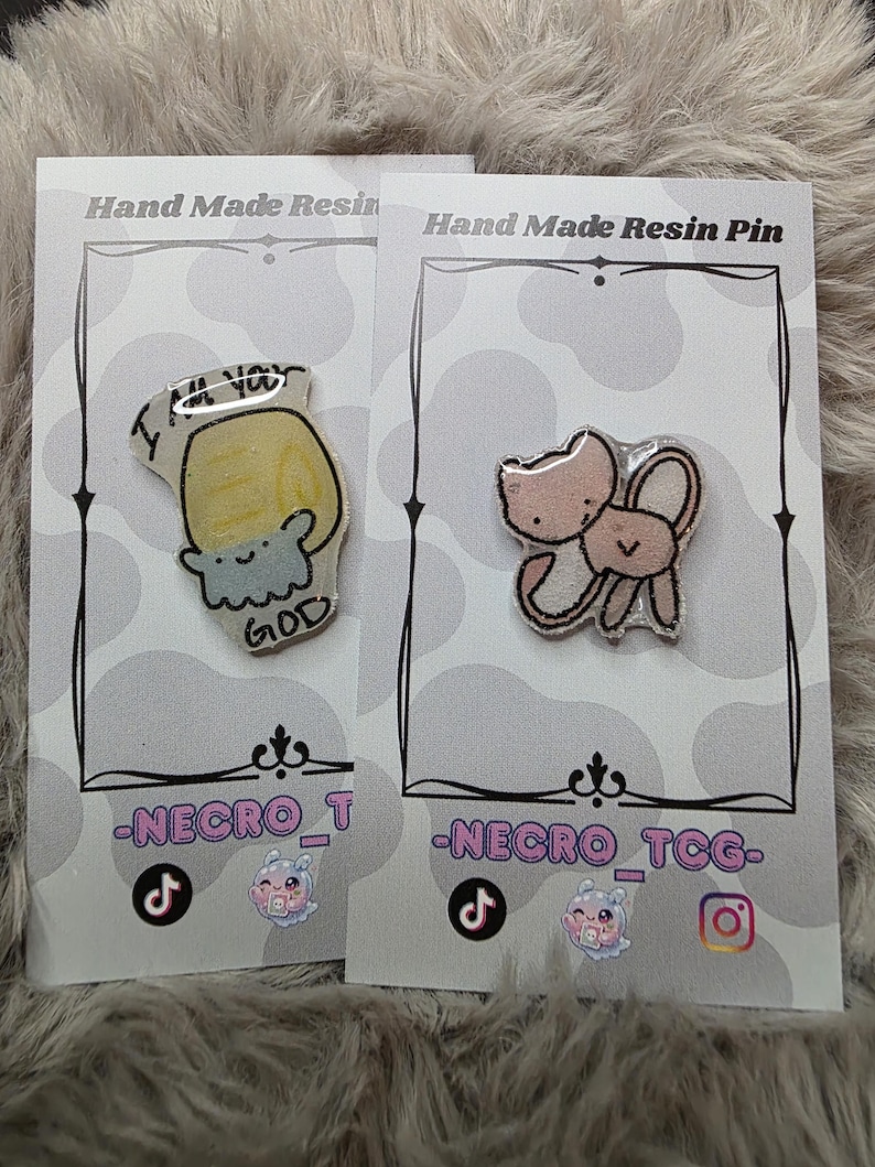 May include: Two handmade resin pins on display. One pin features a yellow design with the text "I'M YOU GOD". The other pin has a pink cat and bunny design. Both pins are on white cards with black borders.