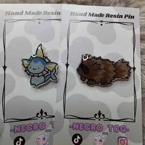 May include: Two handmade resin pins on display. One pin features a blue and white cartoon character with yellow accents. The other pin depicts a brown cartoon raccoon. The pins are on cards with the text "Hand Made Resin Pin" and social media handles.