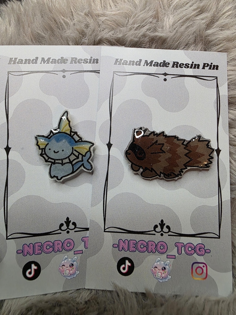 May include: Two handmade resin pins on display. One pin features a blue and white cartoon character with yellow accents. The other pin depicts a brown cartoon raccoon. The pins are on cards with the text "Hand Made Resin Pin" and social media handles.