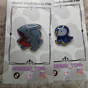 May include: Two resin pins on display. One pin features a gray shark-like creature with a red mouth. The other pin is a blue and yellow penguin. Both pins are on cards that read "Hand Made Resin Pin" and have social media icons.