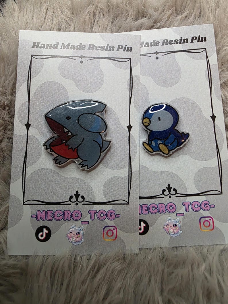 May include: Two resin pins on display. One pin features a gray shark-like creature with a red mouth. The other pin is a blue and yellow penguin. Both pins are on cards that read "Hand Made Resin Pin" and have social media icons.