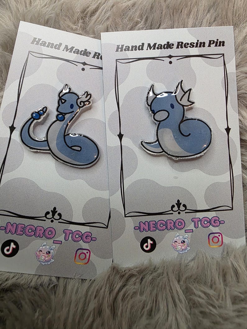 May include: Two handmade resin pins featuring a blue and white dragon design. Each pin is mounted on a gray card with the text "Hand Made Resin Pin" and "-NECRO_TCG-". The pins are on a fluffy, gray surface.