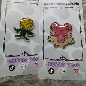 May include: Two resin pins on display cards. One pin is a yellow sunflower with a smiling face and green leaves. The other is a pink creature with wings. The cards read "Hand Made Resin Pin" and feature social media icons.