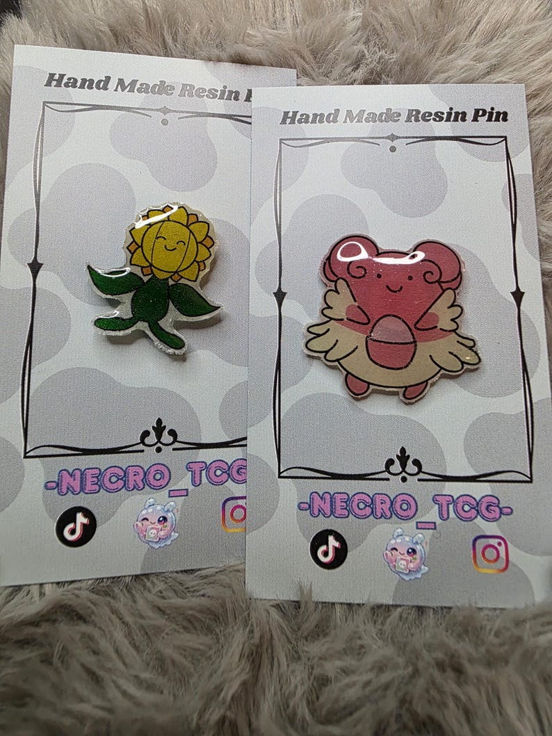 May include: Two resin pins on display cards. One pin is a yellow sunflower with a smiling face and green leaves. The other is a pink creature with wings. The cards read "Hand Made Resin Pin" and feature social media icons.