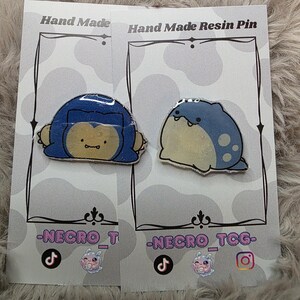 May include: Two handmade resin pins featuring cartoon characters. One pin depicts a blue and yellow character, the other a blue and white character. Each pin is mounted on a gray card with the text "Hand Made Resin Pin" and social media handles.