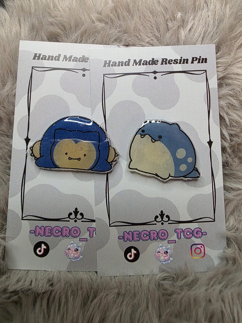 May include: Two handmade resin pins featuring cartoon characters. One pin depicts a blue and yellow character, the other a blue and white character. Each pin is mounted on a gray card with the text "Hand Made Resin Pin" and social media handles.