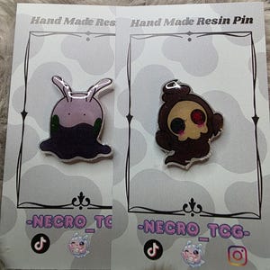 May include: Two resin pins on display cards. One pin is a purple slug-like creature with green accents. The other is a brown skull-like figure with a red eye. The cards read "Hand Made Resin Pin" and feature the text "-NECRO_TCG-".