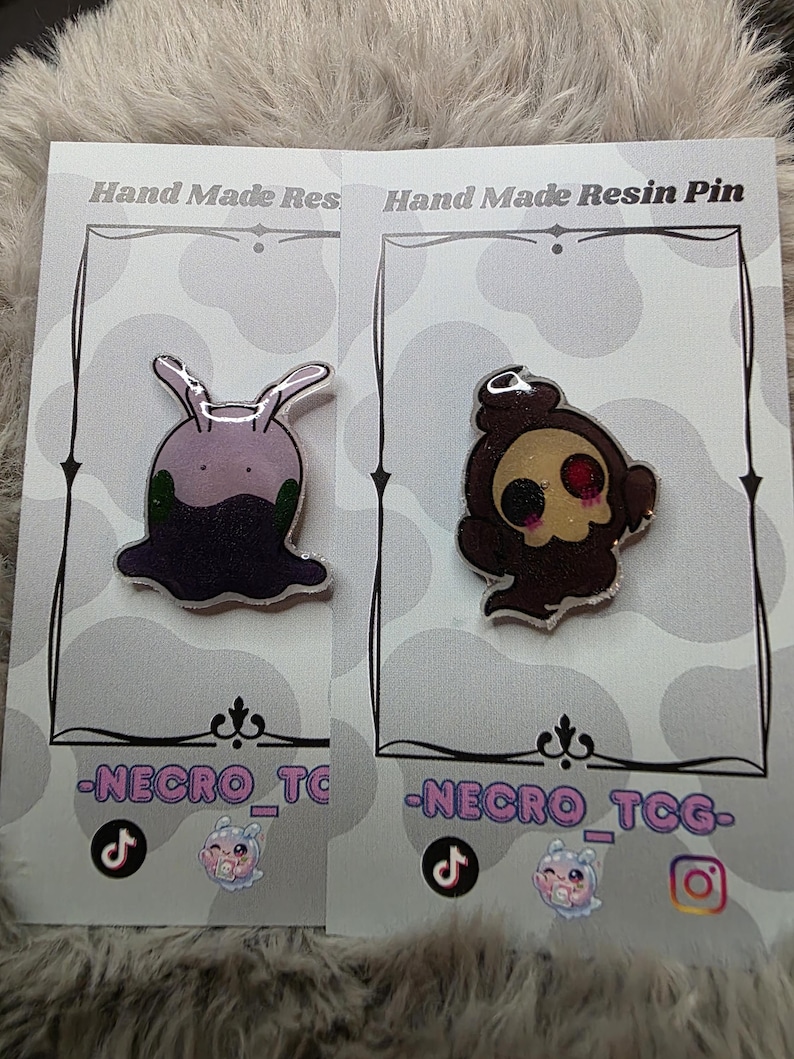 May include: Two resin pins on display cards. One pin is a purple slug-like creature with green accents. The other is a brown skull-like figure with a red eye. The cards read "Hand Made Resin Pin" and feature the text "-NECRO_TCG-".