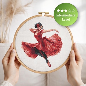 May include: A cross-stitch embroidery hoop featuring a dancer in a red dress. The design is of an intermediate level, as indicated by the green circle with stars. The dancer is in mid-motion, with arms outstretched and a flowing skirt.