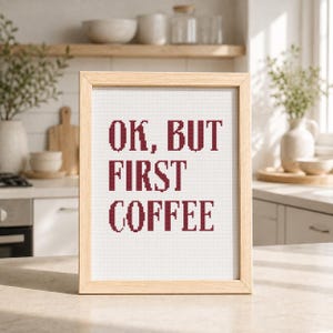 May include: A framed cross-stitch piece featuring the dark red text "OK, BUT FIRST COFFEE" stitched onto a white fabric background. The artwork is enclosed in a light-colored wooden frame with a simple, clean design and a rectangular orientation.