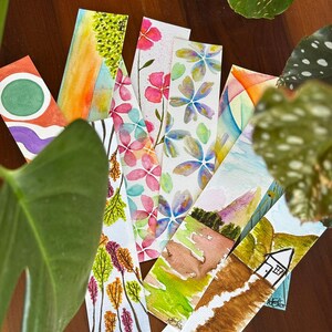 Pode incluir: Uma coleção de marcadores de aquarela com vários designs. Os marcadores apresentam padrões florais, paisagens e arte abstrata em cores vibrantes como rosa, azul, verde e laranja. Cada marcador é retangular e parece ser pintado à mão.