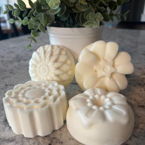 May include: Four ivory-colored, decorative soaps with floral designs. The soaps are arranged on a speckled surface, with a small white pot of greenery in the background. The soaps have different flower-like shapes.