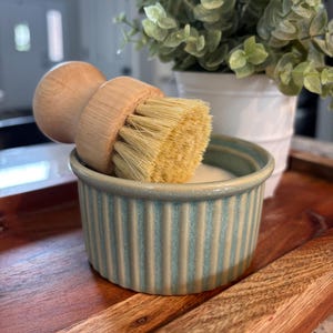 May include: A round wooden dish brush with light-colored natural bristles resting inside a small, round, light blue and white vertically ribbed ceramic ramekin. The brush features a smooth, rounded handle designed for a comfortable grip during use.