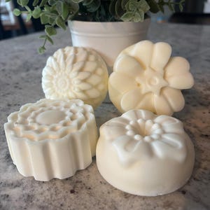 May include: Four off-white, solid, molded soap bars with distinct floral and geometric patterns. Each bar features a unique embossed design, including a sunburst, a multi-petaled flower, a scalloped edge, and a rounded floral shape.