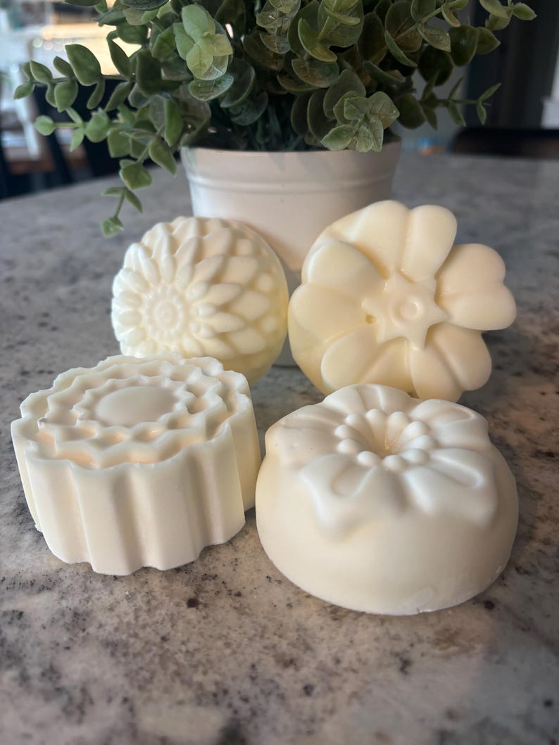 May include: Four off-white, solid, molded soap bars with distinct floral and geometric patterns. Each bar features a unique embossed design, including a sunburst, a multi-petaled flower, a scalloped edge, and a rounded floral shape.
