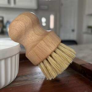May include: A round wooden dish brush with a light-colored, natural wood handle and stiff, light beige bristles. The handle features a contoured, ergonomic shape designed for a firm grip, while the dense bristles are arranged in a circular pattern.