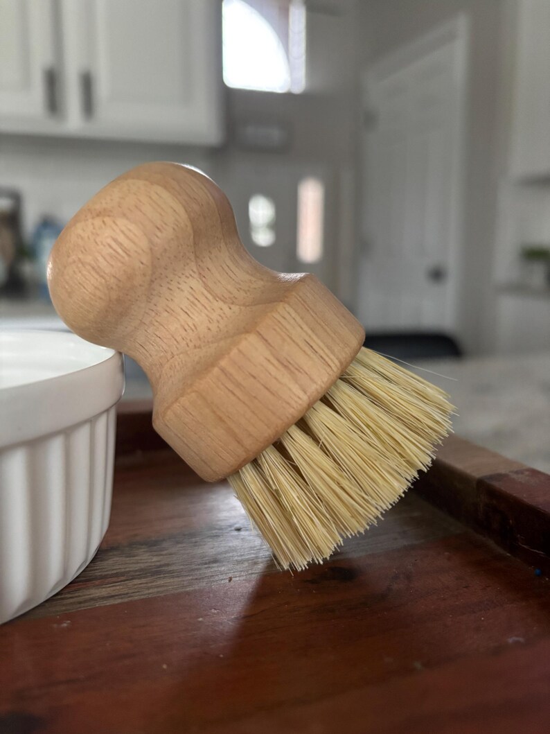 May include: A round wooden dish brush with a light-colored, natural wood handle and stiff, light beige bristles. The handle features a contoured, ergonomic shape designed for a firm grip, while the dense bristles are arranged in a circular pattern.